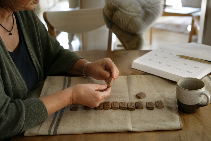 What are runes: Elder Futhark tiles on a kitchen table Hands turning over a carved wooden rune tile beside an open reference book on a kitchen table