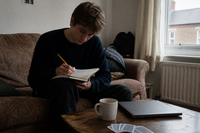 Young man on a sofa writing predictions in a small notebook, playing cards face down on the coffee table
