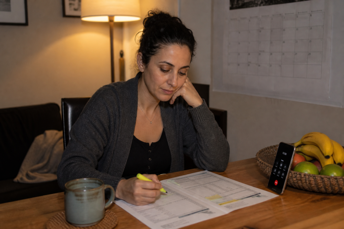 Woman at a dining table checking a printed bank statement with a highlighter, phone propped nearby