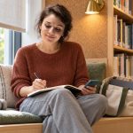 What Is an Empath? Sensitivity, Spiritual Meaning and Everyday Support A woman with a wavy brown bob writes in a notebook on a library window bench, with a teal phone and headphones nearby.