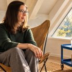 What Qualities and Abilities Does an Empath Have? A woman with straight brown hair and glasses sits by a triangular window, with a closed notebook and folding phone nearby.
