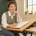 The Four Clairs: Spiritual Meanings and Optional Reflection Exercises A short-haired woman in a sage vest writes in a notebook at a slanted drawing desk.