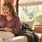 Connecting with Your Higher Self: Five Reflection Questions Woman writing in a notebook on a cream window seat, with a purple phone on a small sage side table.