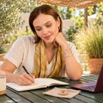 Finding Balance in Mind, Body and Spirit: A Four-Area Weekly Review Woman writing in a notebook on a garden terrace beside a laptop and a wooden Gebo rune