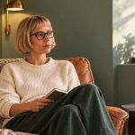 Past-Life Bonds: Three Lenses for Meaning and Choice Woman with a blonde bob and glasses sitting with a closed notebook in a leather chair beside a window.