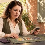 Are Psychics Real? A woman in a green waistcoat holds a tarot deck beside three cards on a round table in a shaded patio.