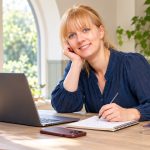 Fake vs Real Psychics and Mediums: How to Tell an Honest Reader From a Fraud A woman with strawberry-blonde hair reviews notebook notes beside a laptop, phone, oracle deck and small amethyst.