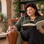 The History of Runes: What Scholars Know, What Was Lost, and What Is Modern Woman reading a history book in a leather armchair beside wooden Isa and Gebo study tiles