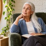 Rune Reflection Without Body Placement: An Optional Exercise Silver-haired woman with an open notebook beside a wooden Isa rune on a small stool