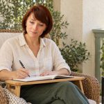 Spirit-Guide Journaling: Five Claims and an Optional Reflection Method An auburn-haired woman writes in a notebook on a bamboo lap desk in a rattan chair beside a tablet.
